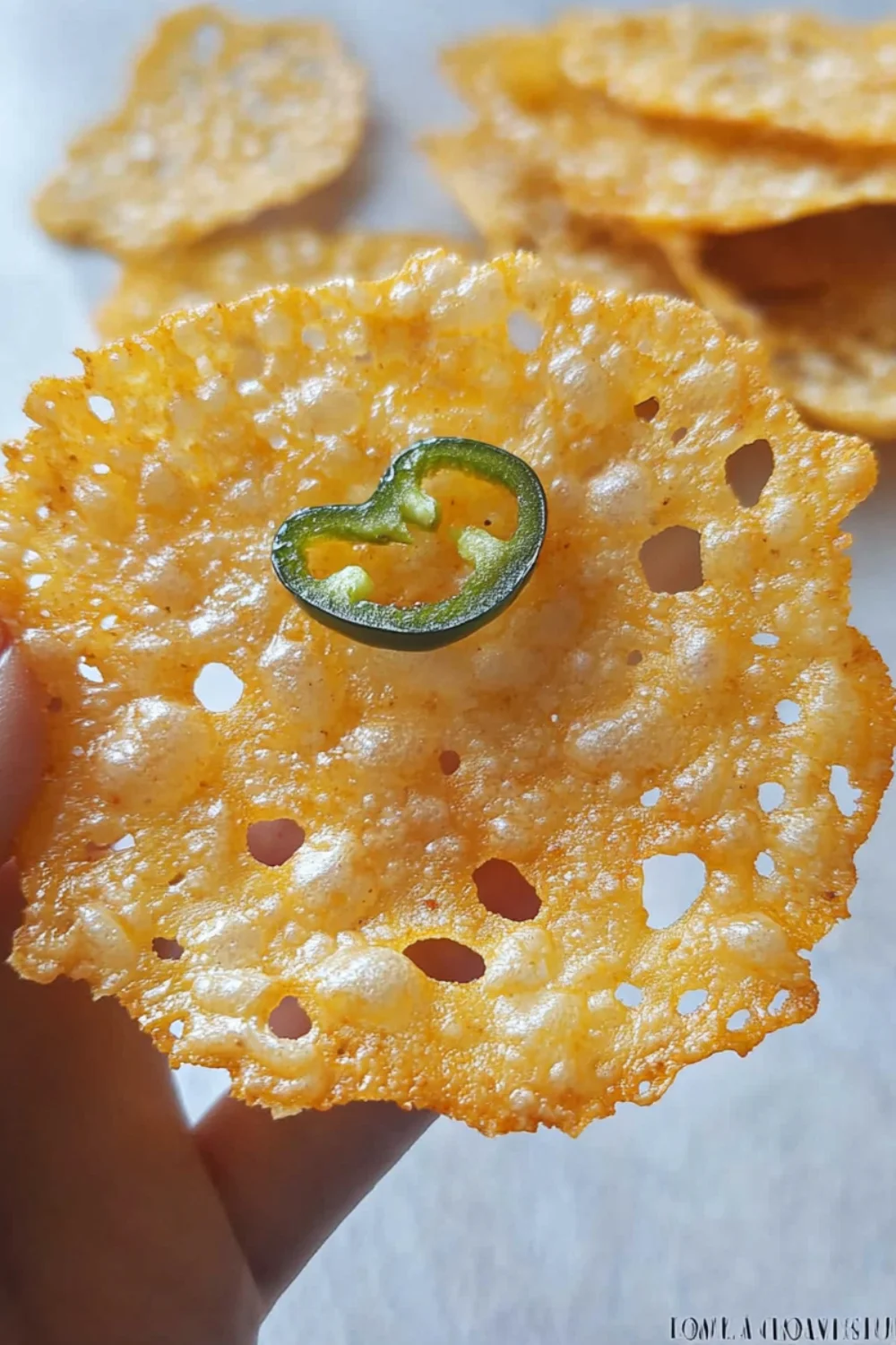 Eine Hand hält einen knusprigen Käsechip mit einer Scheibe Jalapeño in der Mitte.